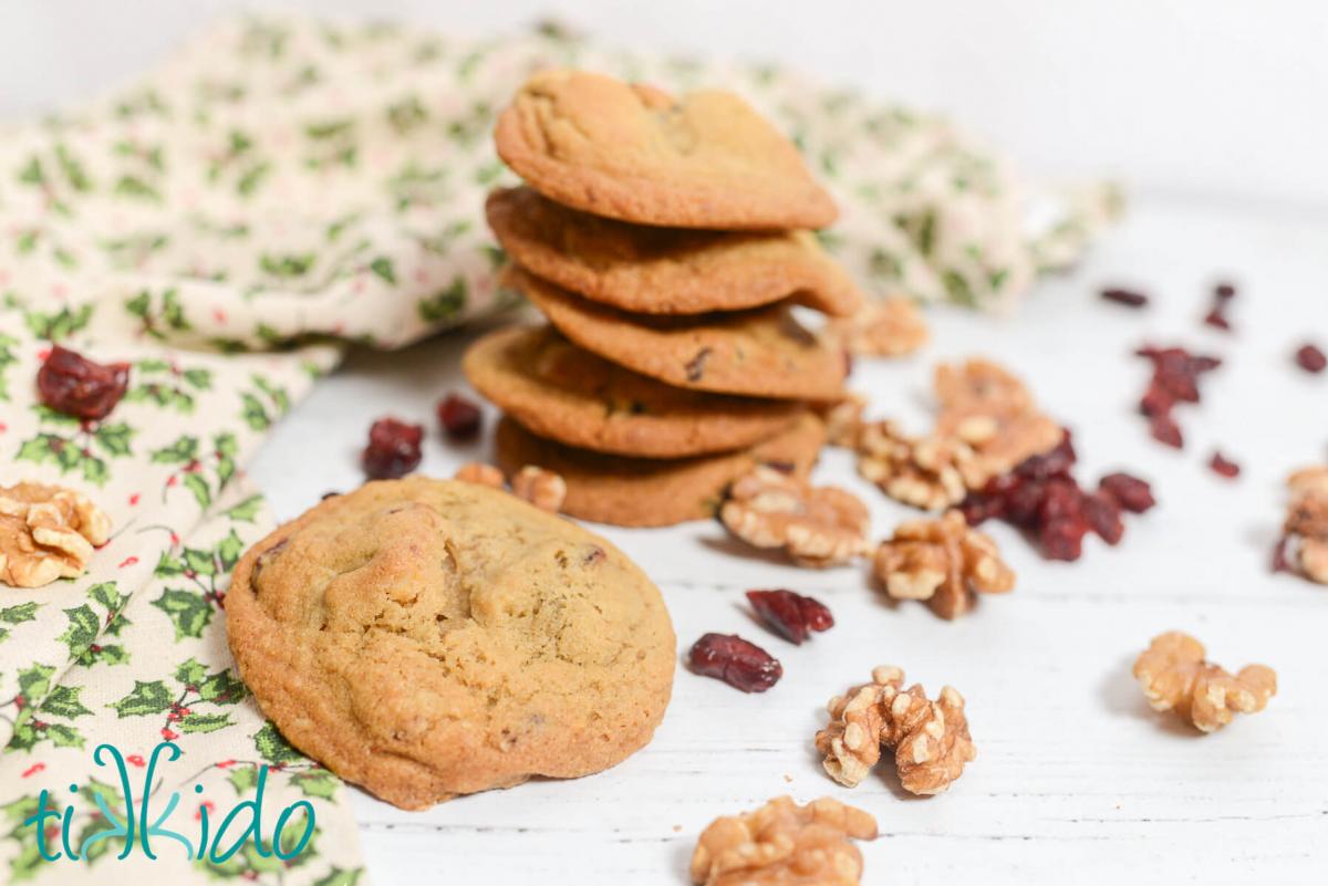 Stack of Cranberry Walnut Cookies surrounded by walnut halves and dried cranberries and a holly print Christmas tea towel on a white wooden surface.
