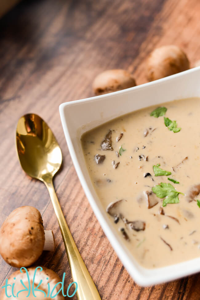 Homemade cream of mushroom soup in a white bowl, next to a golden spoon and fresh mushrooms.