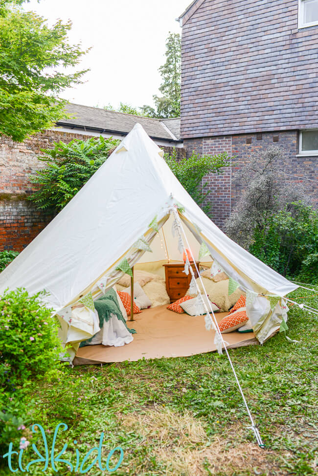 Bell tent set up in a backyard, with bunting decorating the exterior, and a bed, two beanbag chairs, a small table, and many turquoise, white, and peach pillows scattered on the floor of the tent.