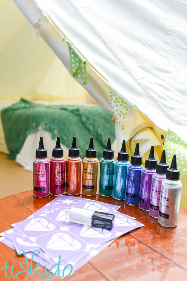 Bottles of body glitter arranged in the order of the rainbow, with glitter tattoo stencils and glue in front of the bottles, on a wooden table.