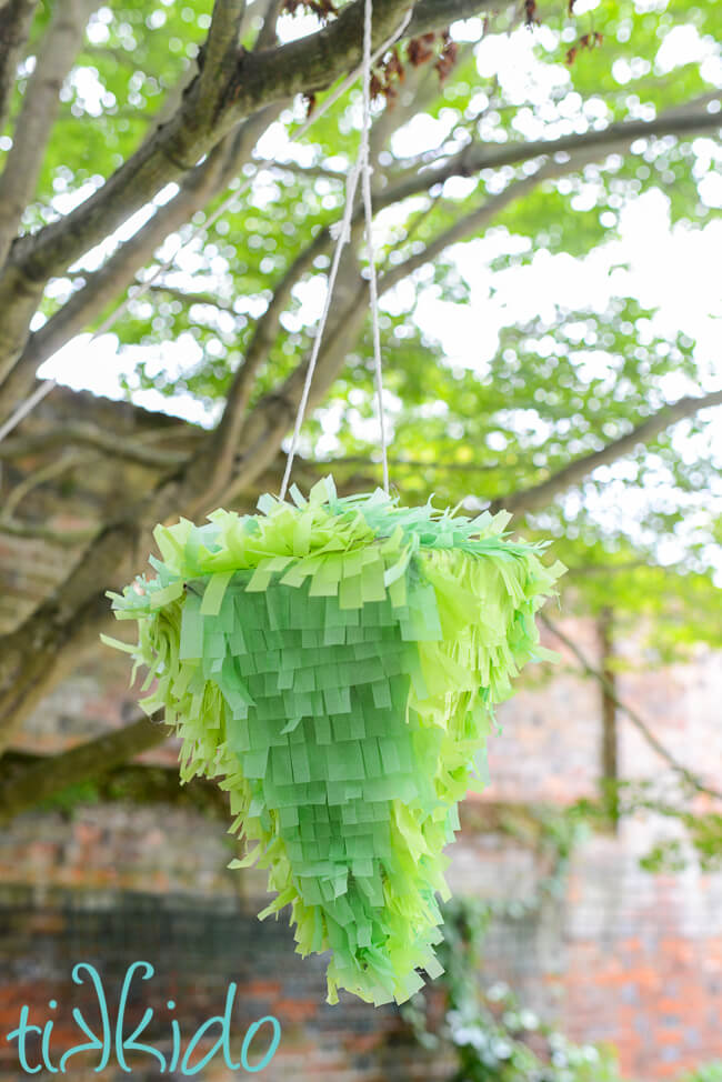 Pinata that looks like a green crystal hanging from a tree in front of a brick wall.
