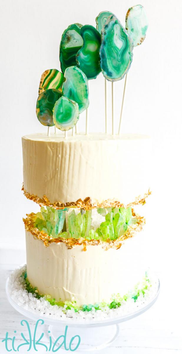 Birthday cake for a Crystal Themed Birthday Party.  The cake topper is made from 9 green agate slices on white lollipop sticks. The cake is two tiers of the same size, covered in white Italian meringue buttercream, and there's space between the two layers, where you can see green sugar crystals "growing" out of the top and bottom cake, like a cave or a geode.