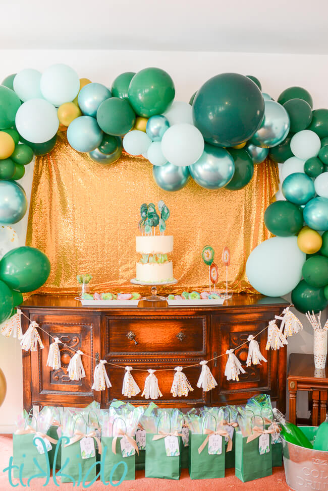 Dessert table at the Crystal Themed Birthday Party, with a sparkling gold background, green and gold balloon garland, tissue paper tassel garland, and green gift bags set in front of a wooden sideboard.  On the sideboard there's a cake decorated with green crystals, lollipops that look like agate slices, sugar cookies that look like geodes, and green and gold rock candy.