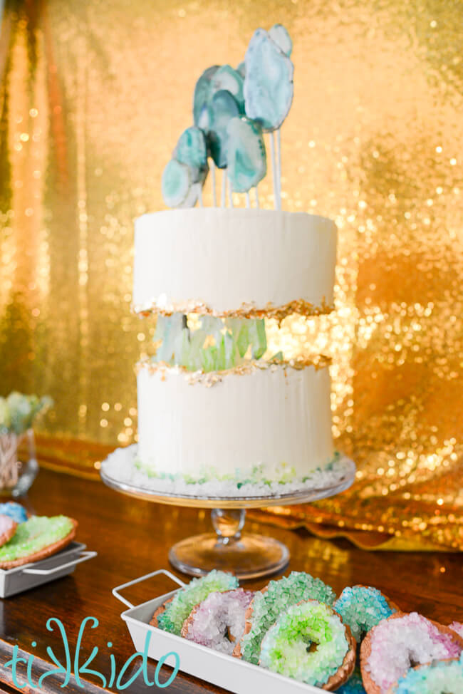 White rectangular tray of sugar cookies that look like geodes, in greens, blues, and purples.  A crystal geode cake and gold sequin backdrop are blurred in the background.