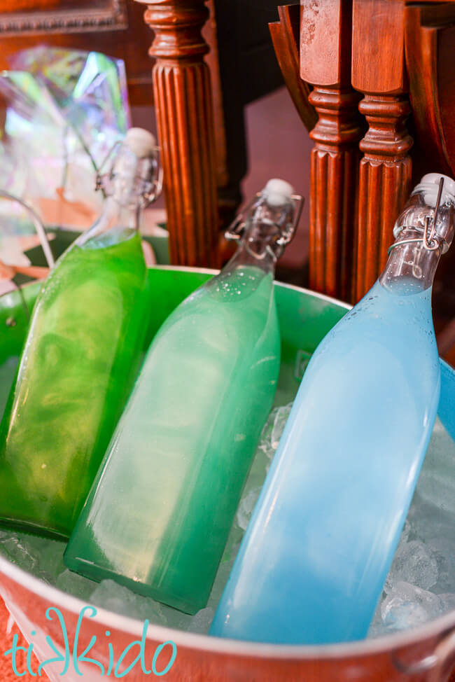 Three bottles of koolaid shimmering and looking like magical potions, nestled in an ice filled drinks bucket.  The bottle on the left is filled with shimmering green liquid, the middle bottle with turquoise punch, and the bottle on the right with blue koolaid.