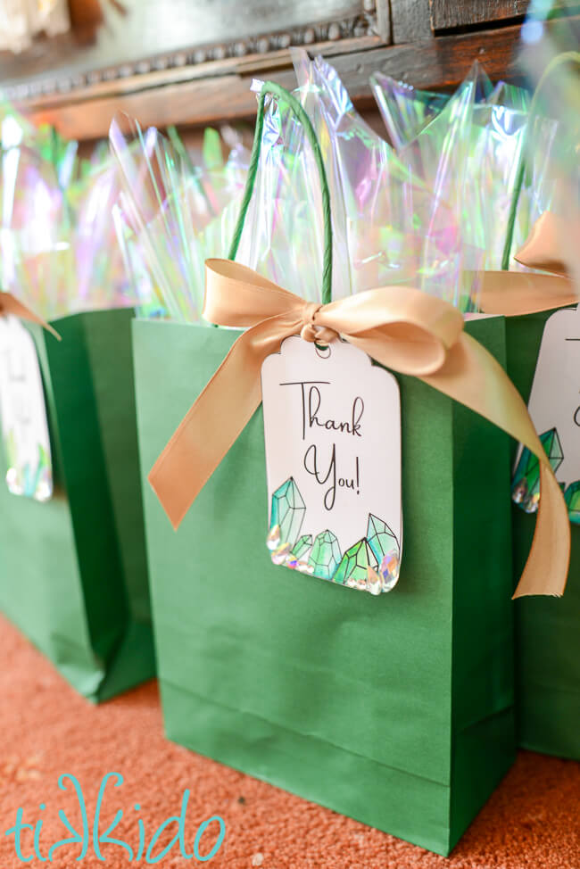 Green gift bags embellished with a printable crystal gift tag that says "Thank You," attached to the bag with a gold satin ribbon tied in a bow.  Iridescent  cellophane peeks out of the top of the gift bag.