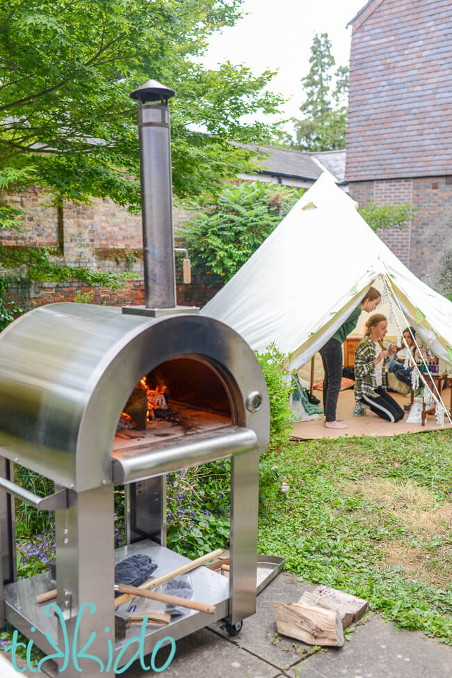 Back Garden with a Stainless steel pizza oven with a fire in the foreground, and a white bell tent decorated in boho style in the background.