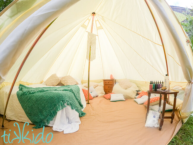 Wide interior shot of a bell tent set up for glamping, with a full bed, two beanbag chairs, two small wooden tables, and many pillows scattered around the floor.  The closest small table is set up with materials for making glitter tattoos.