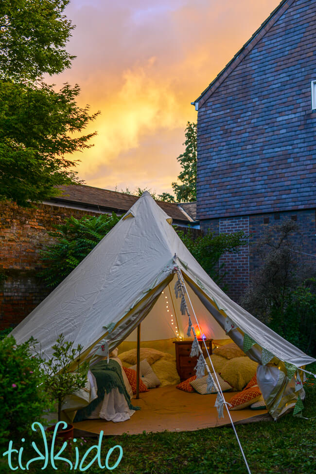 View of a bell tent in a backyard set up for glamping at sunset, with fairy lights glowing inside the tent and a beautiful orange sunset in the sky behind.