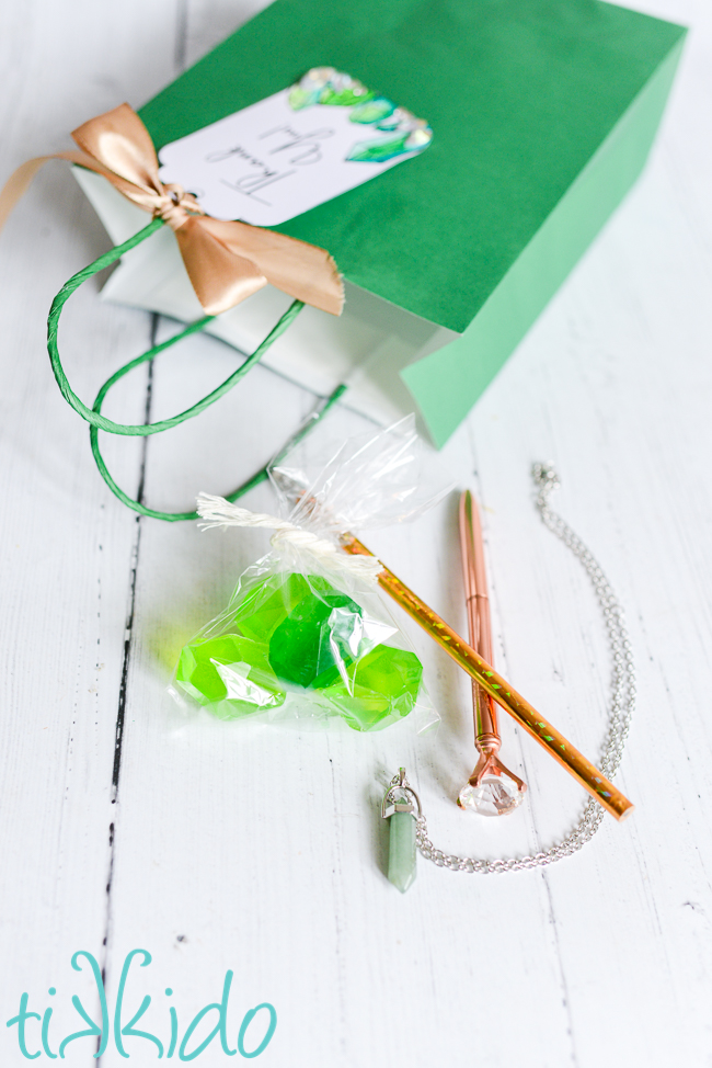 Gift bag at the Crystal Birthday Party on its side on a white wooden surface, with the contents of the bag in front of it:  a clear bag with four green hand soaps shaped like gemstones, a gold shiny pencil, a rose gold pen topped with a large artificial diamond, and a necklace with a pale green crystal pendant and silver chain.
