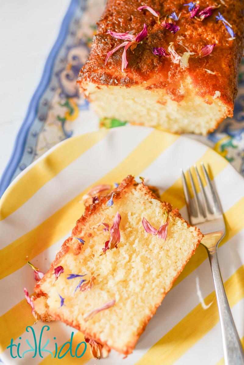 Slice of lemon drizzle cake decorated with dried flower petals, on a yellow and white striped plate with a fork next to the cake.  You can just see the sliced loaf of lemon drizzle cake in the background.