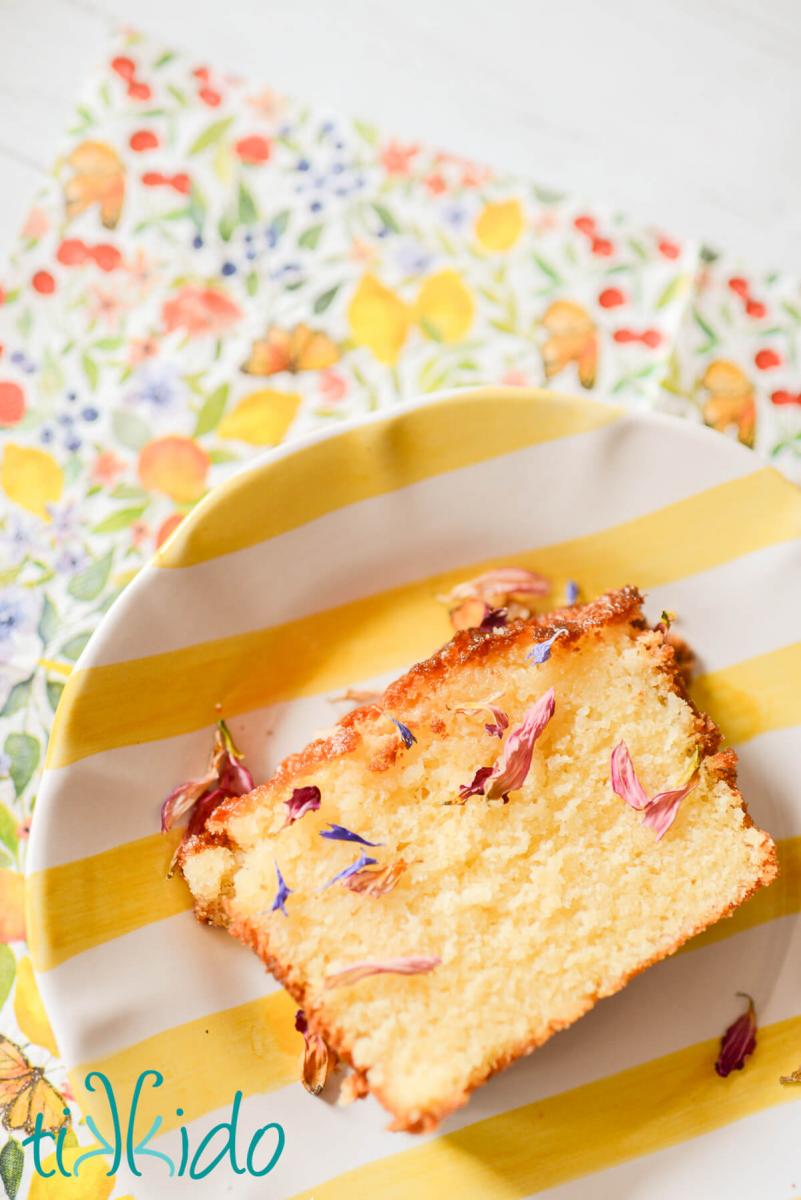 Slice of lemon drizzle cake decorated with homemade dried edible flower petals, on a yellow and white striped plate, on top of a floral print napkin.