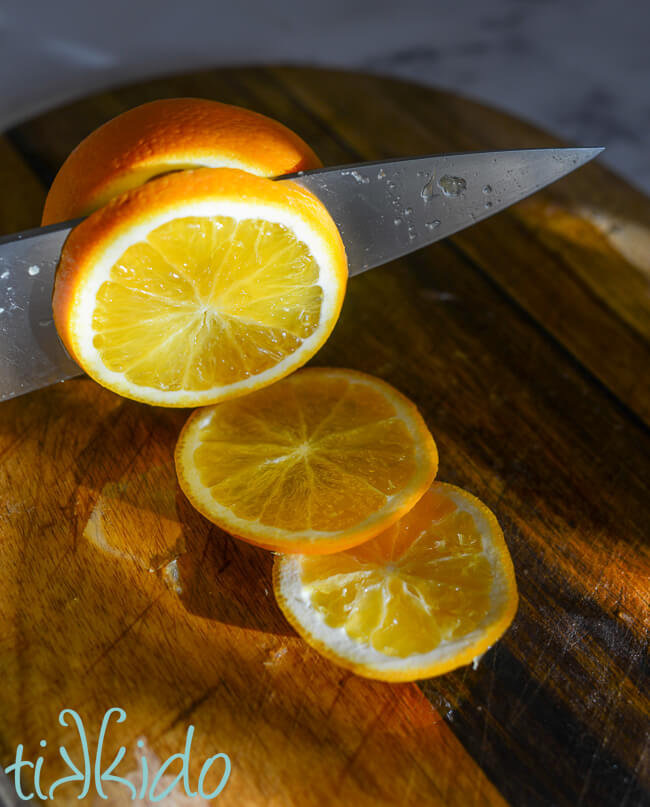 Fresh orange being cut into thin slices on a wooden cutting board.