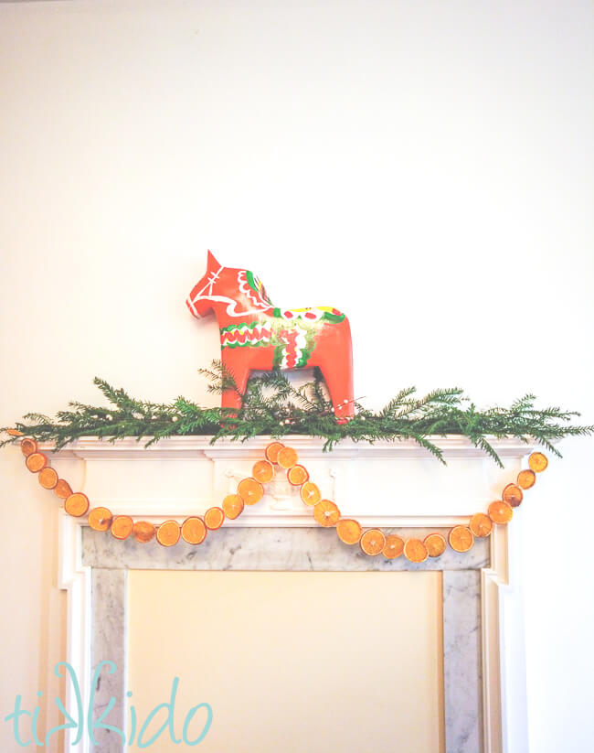 Dried Orange Garland hung from a fireplace mantel.  The mantel is also decorated with fresh evergreen branches and a large dala horse.