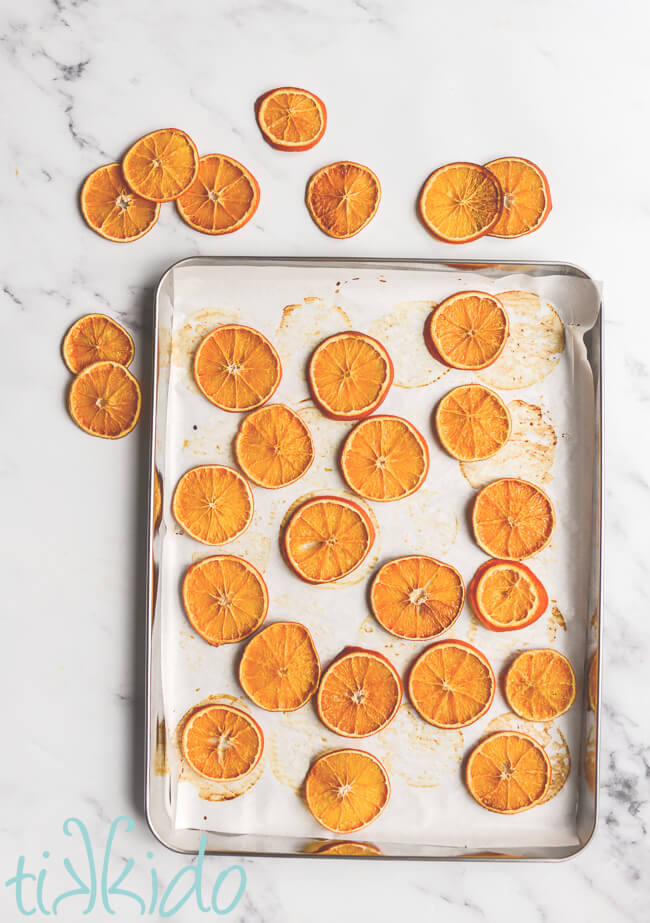 Dehydrated orange slices on a parchment lined baking sheet on a white marble surface.