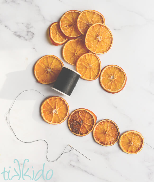 Slices of dried oranges and a spool of black thread on a white marble surface.  Four orange slices have been strung together with a needle and thread to make an orange garland.