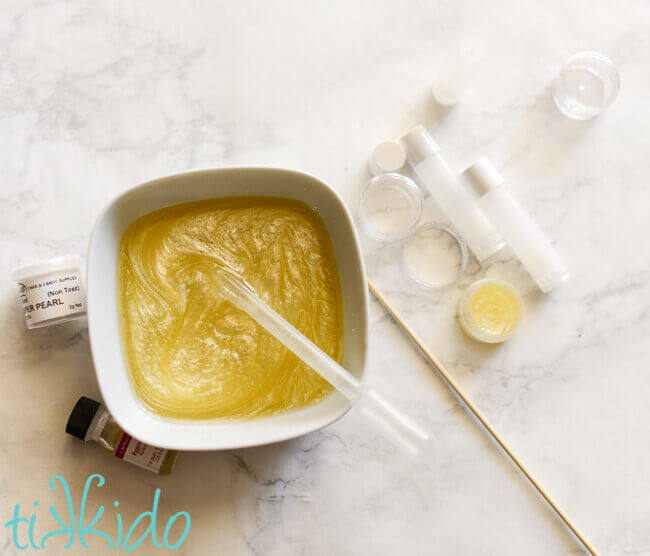 Shimmering homemade lip balm being made in a white bowl, with a pipette being used to transfer mixture to a lip balm container.