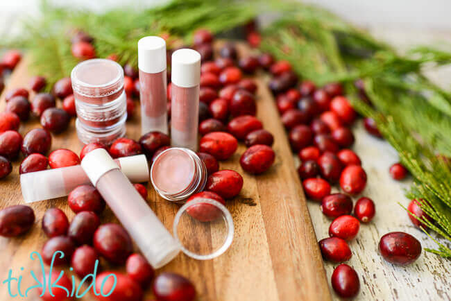 Pots and twist up containers of homemade cranberry lip gloss surrounded by fresh cranberries and evergreen branches.
