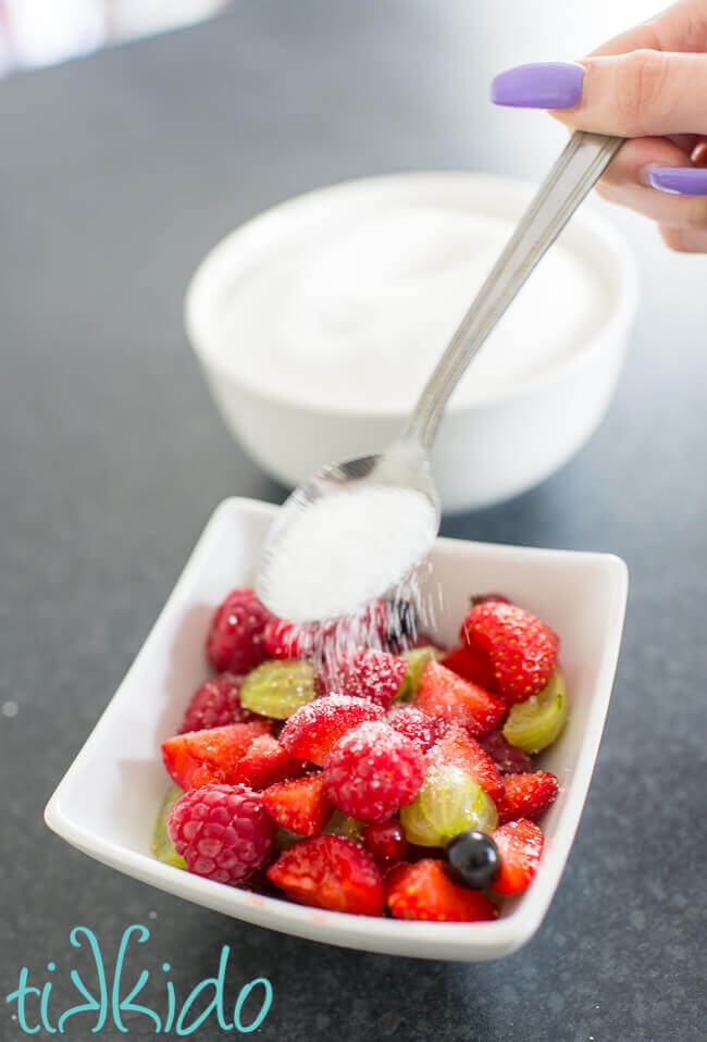 Sugar being sprinkled on a bowl full of fresh English summer berries, including strawberries, gooseberries, currants, and raspberries.