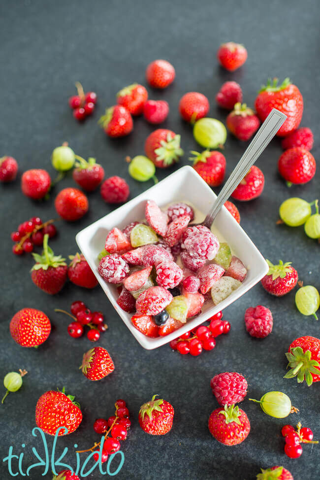 Summer berries and cream in a white bowl, on a black background, surrounded by fresh summer berries.