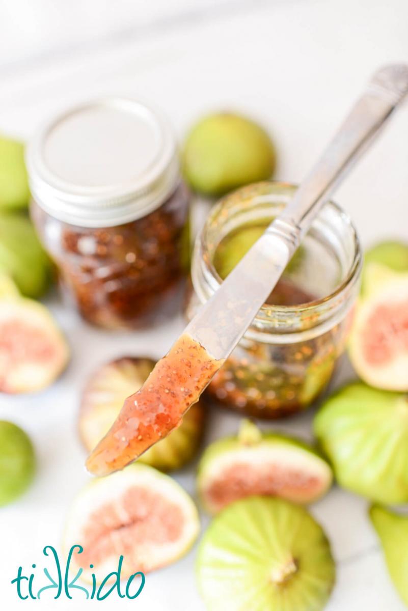 Butter knife coated in homemade fig jam resting on top of a jar full of fig jam, surrounded by fresh figs.