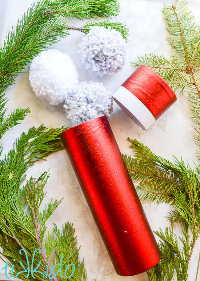 Yarn pom poms spilling out of a cylindrical wine bottle gift box for a DIY Pom Pom Snowball Fight Kit