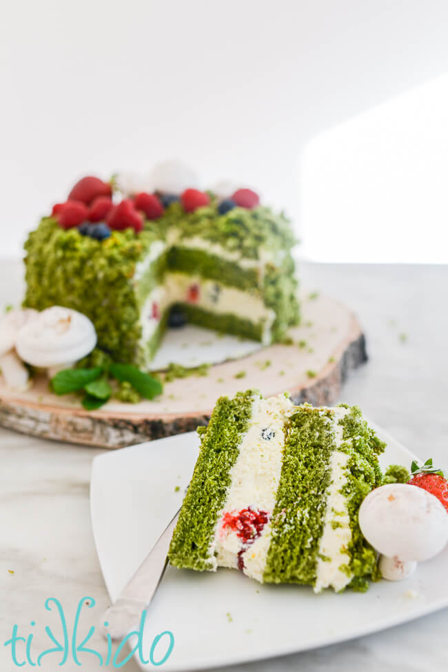 Slice of green moss cake (made with spinach, not moss) in front of the rest of the cake, which is decorated to look like it is covered in moss, berries, and mushrooms (made from meringue).