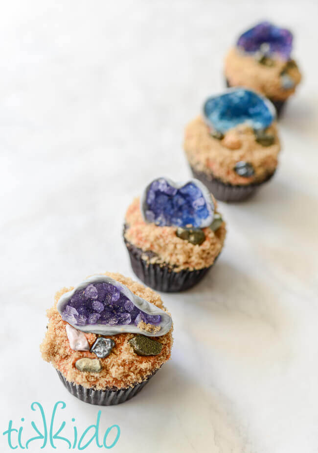 Line of cupcakes with edible gum paste and sugar crystal geode cupcake toppers on a white marble surface.