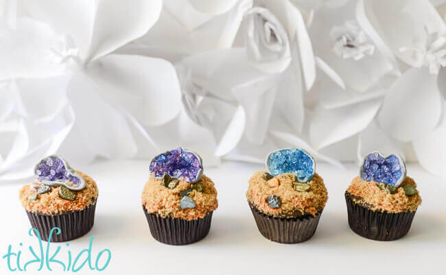 line of four cupcake topped with edible gum paste and sugar crystal geode cupcake toppers on a white background.