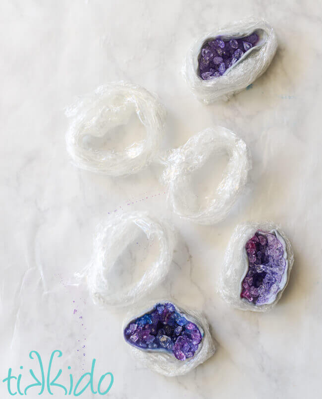 Three purple and blue rock candy edible geode cupcake toppers drying in cling wrap nests on a white marble background.