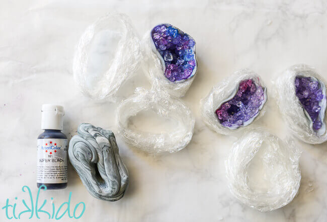 Gel food coloring, marbled grey gum paste, nests of cling wrap, and three edible geode cupcake toppers drying on a white marble surface.
