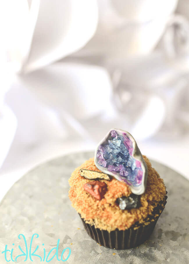 Cupcake topped with brown sugar dirt, chocolate rocks, and a gum paste and rock candy edible geode cupcake topper.