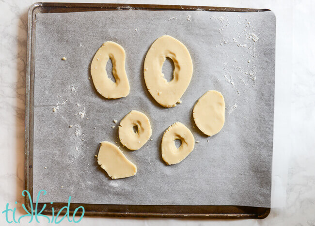 Sugar cookie dough cut into 6 oblong, irregular shapes, four of which have a hole in the center, on a parchment sheet on a cookie sheet.