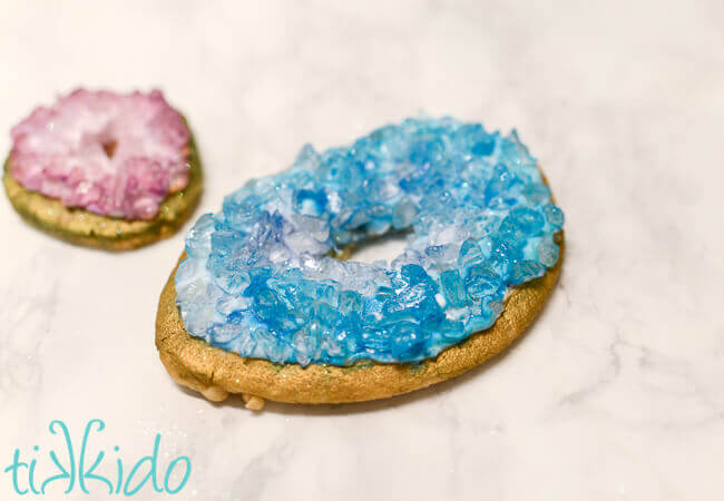 One large blue crystal geode sugar cookie, and one smaller purple geode sugar cookie to its left, both on white marble surface.
