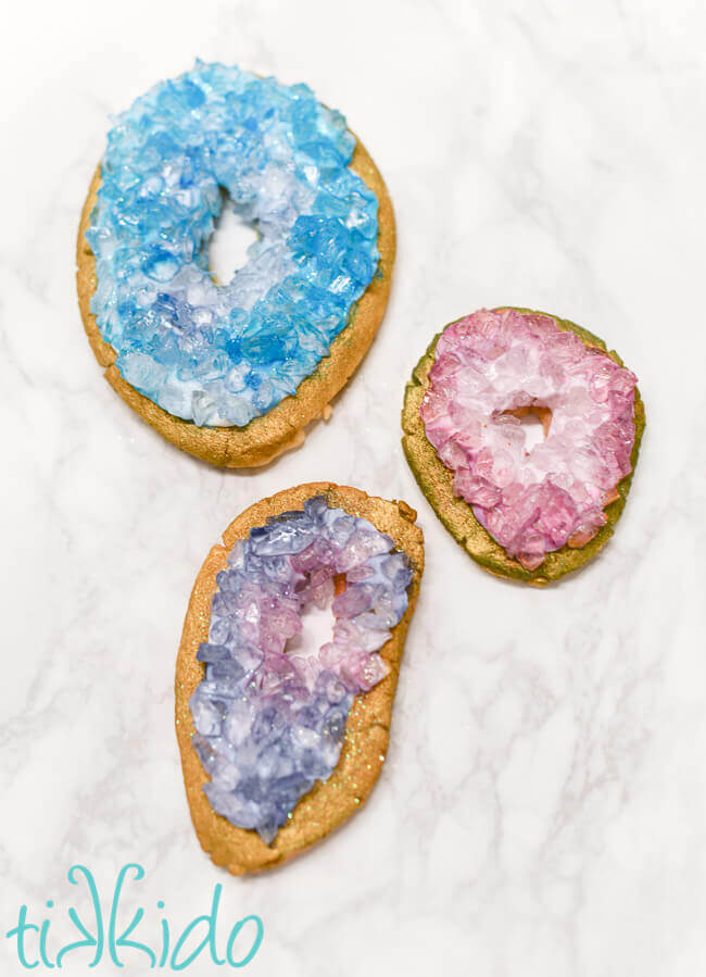 Three geode sugar cookies made with painted rock sugar crystals, one blue and two purple, on a white marble surface.