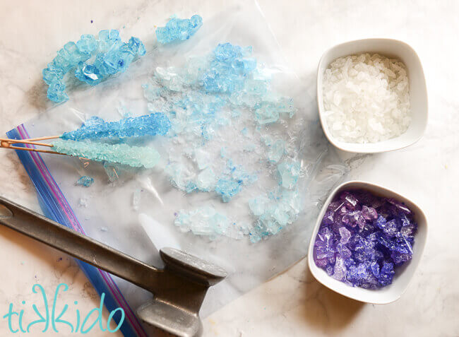 Crushed blue rock candy in a ziplock bag, metal meat mallet on top of the bag, two white bowls with crushed clear and purple rock candy crystals to the right.