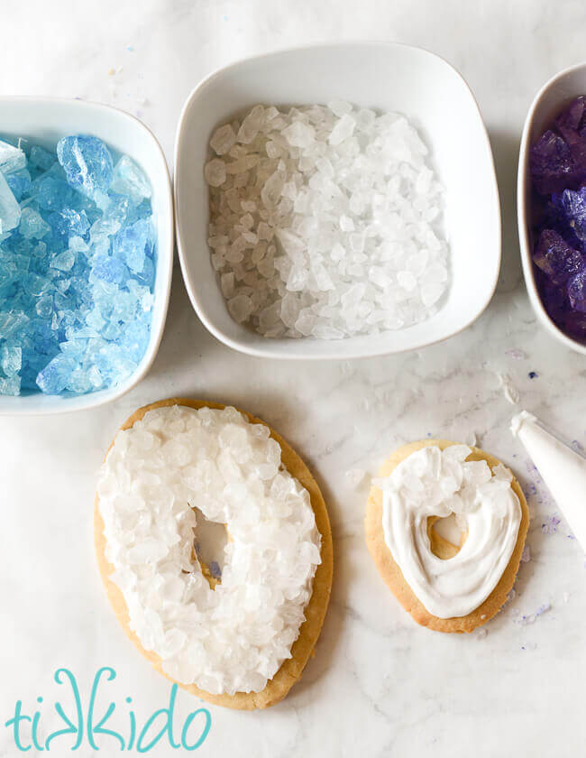 Bowls of rock candy crystals above two sugar cookies iced with white royal icing and clear sugar crystals embedded in the icing.