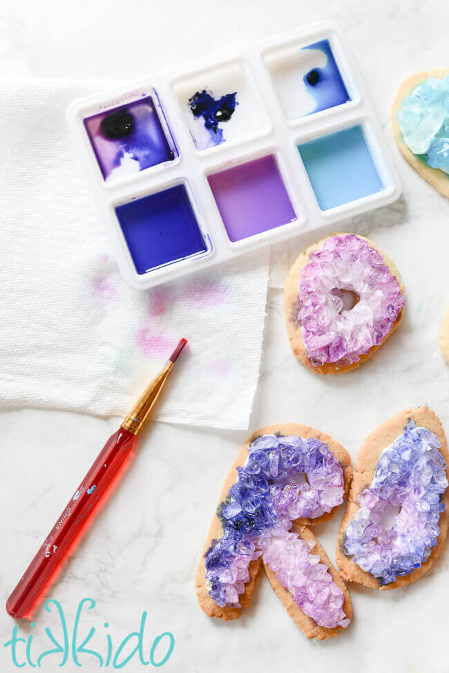 watercolor painting palette with blue and purple food coloring being used as paint, paintbrush, and three painted geode cookies