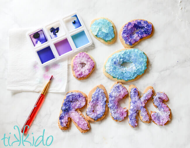 watercolor painting palette with blue and purple food coloring being used as paint, paintbrush, and 9 painted geode cookies