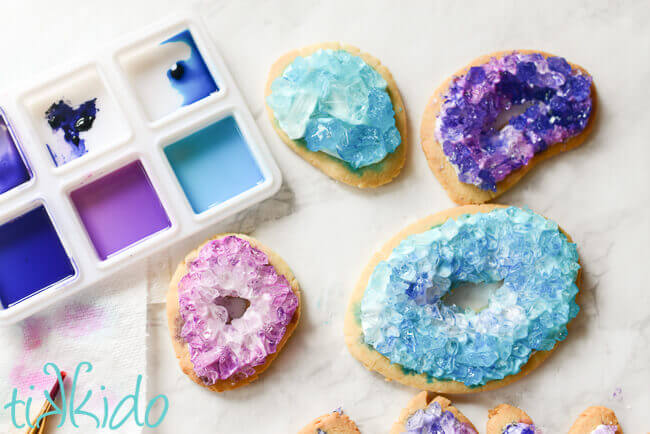 watercolor painting palette with food coloring and four painted geode sugar cookies, two blue, two purple.