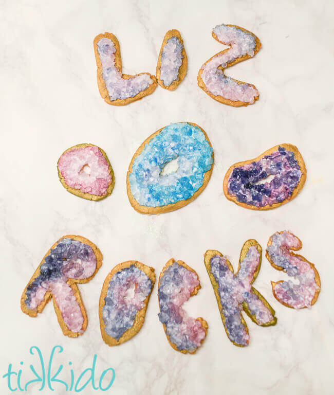 turquoise and purple geode sugar cookies spelling out "Liz Rocks" on a white marble surface.