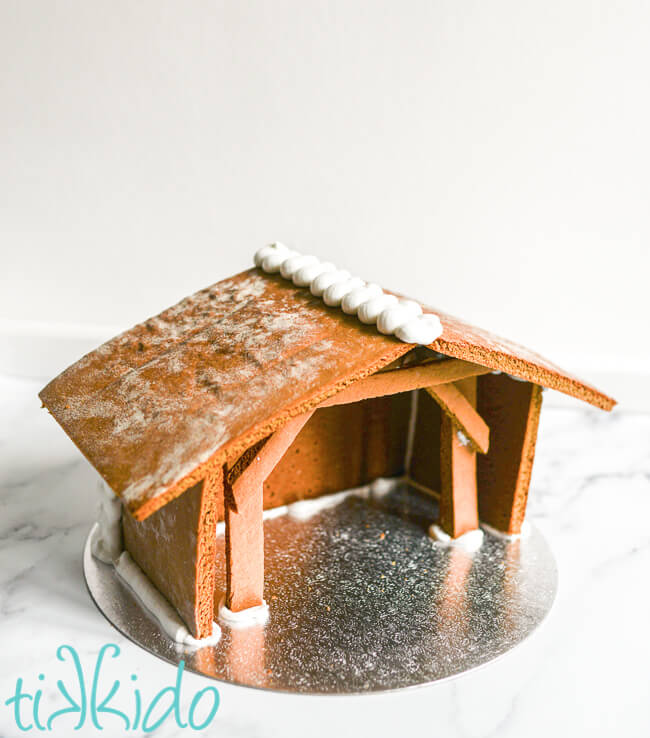 Finished assembled gingerbread house nativity on a silver cake round on a white background.