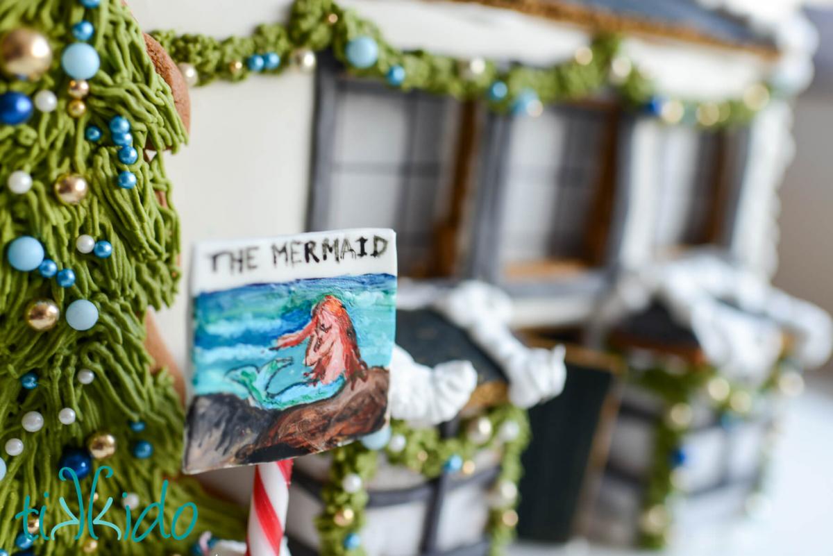 Closeup of the sign for the gingerbread version of The Mermaid pub, painted in food coloring on gum paste, with a candy cane pole.