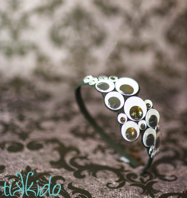 Halloween headband covered in googly eyes standing on a grey damask backdrop.