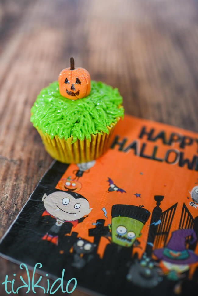 Cupcake on a Halloween napkin, decorated with icing grass and a gum paste pumpkin jack o lantern.