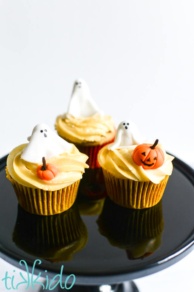three cupcakes decorated with Ghost Cupcake Toppers and small gum paste pumpkins on a black cake plate.