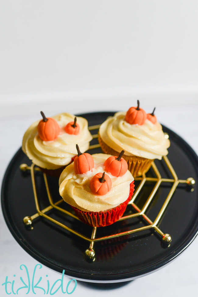 Three cupcakes topped with gum paste pumpkins, on a black cake stand.