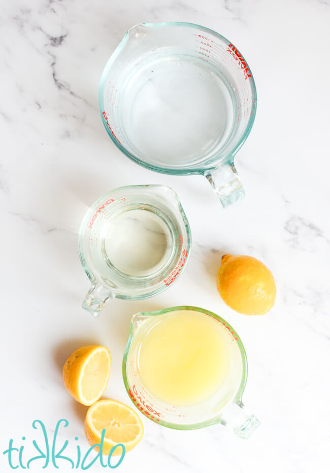 Ingredients for homemade lemonade recipe in measuring cups on a white marble surface, next to two fresh lemons.