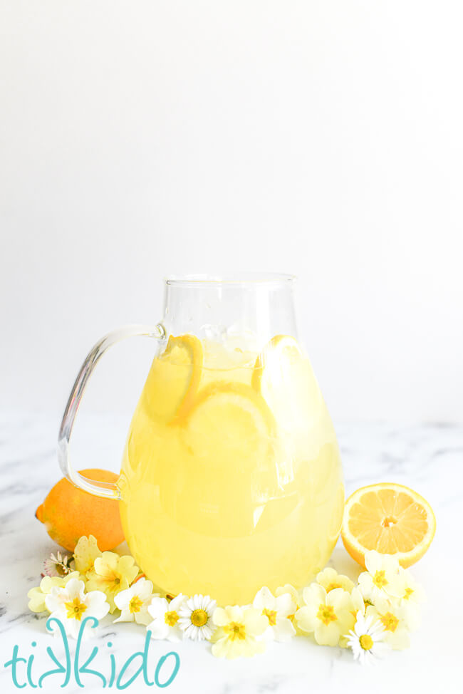Pitcher of homemade lemonade surrounded by white and yellow flowers and two lemons.