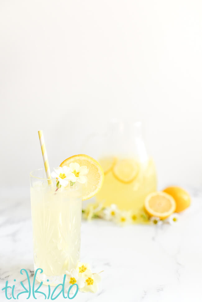 Glass of homemade lemonade garnished with edible yellow and white flowers and a slice of lemon and a yellow and white striped straw.  A pitcher of lemonade and some whole lemons are in the background.